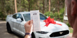 A man holds a winning Lotto ticket in front of a new car.