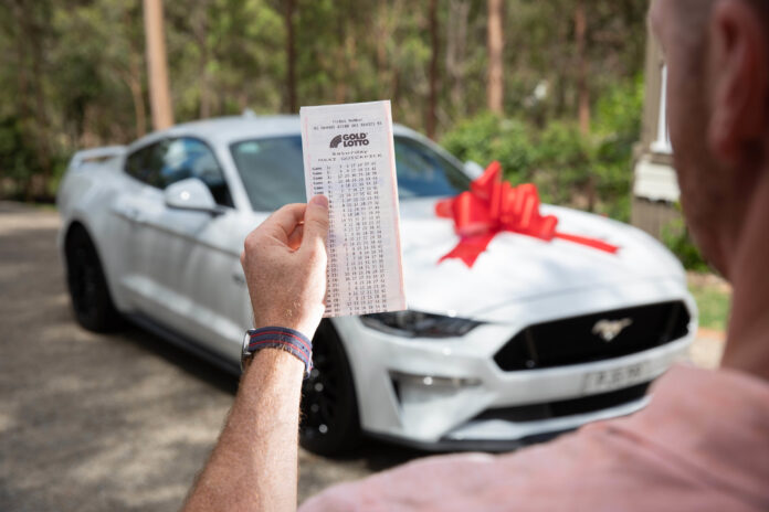 A man holds a winning Lotto ticket in front of a new car.