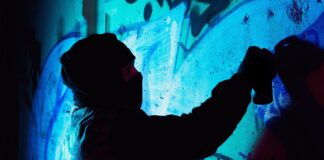 A young hooligan with a spray can stands against a concrete wall with graffiti paintings.
