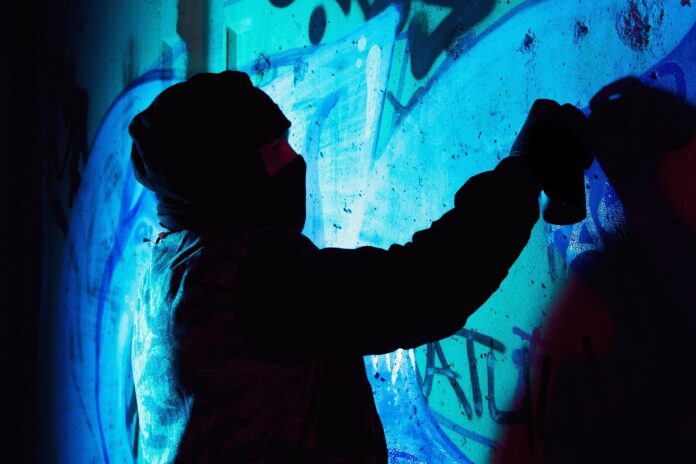A young hooligan with a spray can stands against a concrete wall with graffiti paintings.