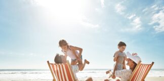 A couple sit in deck chairs on the beach, holding their children, under a clear and bright sky. Picture: Shutterstock.