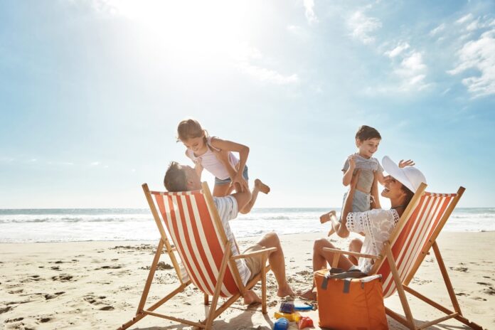 A couple sit in deck chairs on the beach, holding their children, under a clear and bright sky. Picture: Shutterstock.