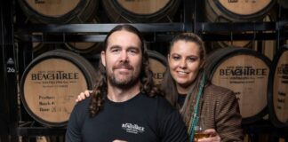 Steve Grace and Kirra Daley of Beachtree Distilling Co hold drinks and stand in front of barrels.