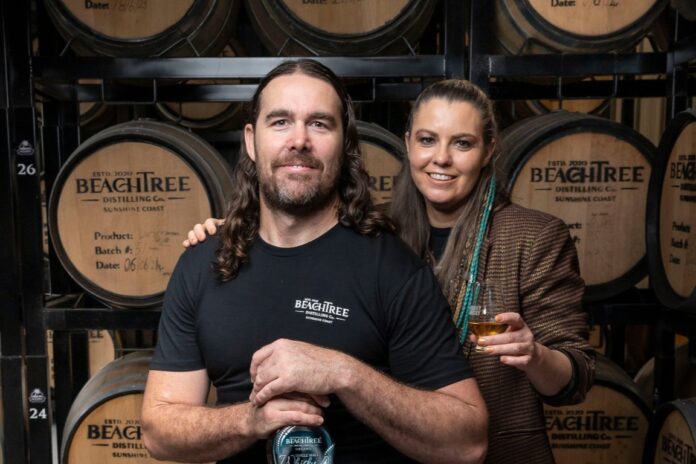 Steve Grace and Kirra Daley of Beachtree Distilling Co hold drinks and stand in front of barrels.