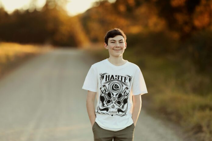 A teen boy wearing a white shirt is standing ona dirt road, smiling.