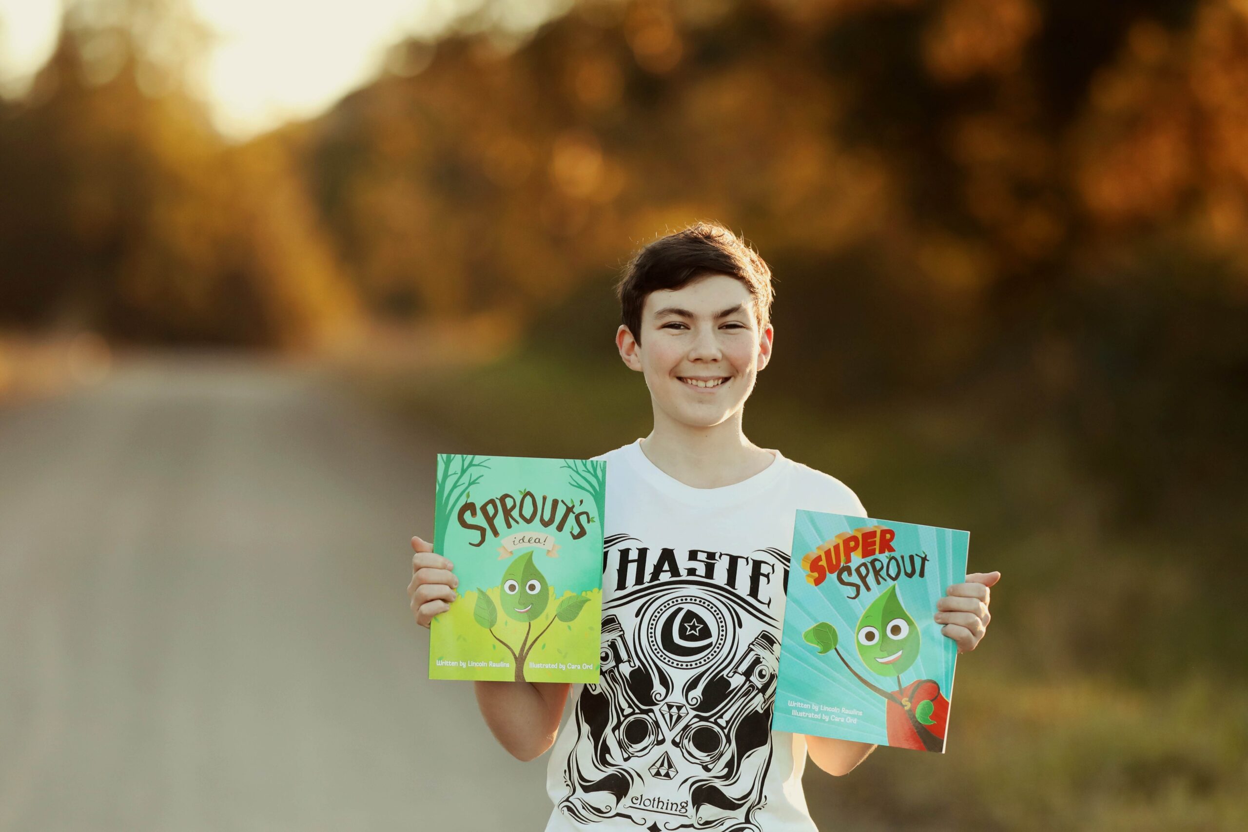 Teenage boy standing on a dirt road while smiling and holding two books.