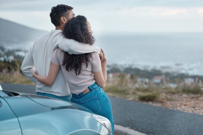A couple standing beside a parked car, looking over the ocean.
