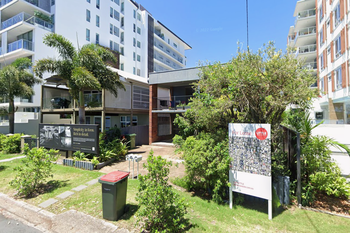 Two-storey brick residential building with a flat roof and front balconies, surrounded by palm trees and shrubs, with taller apartment buildings on either side.