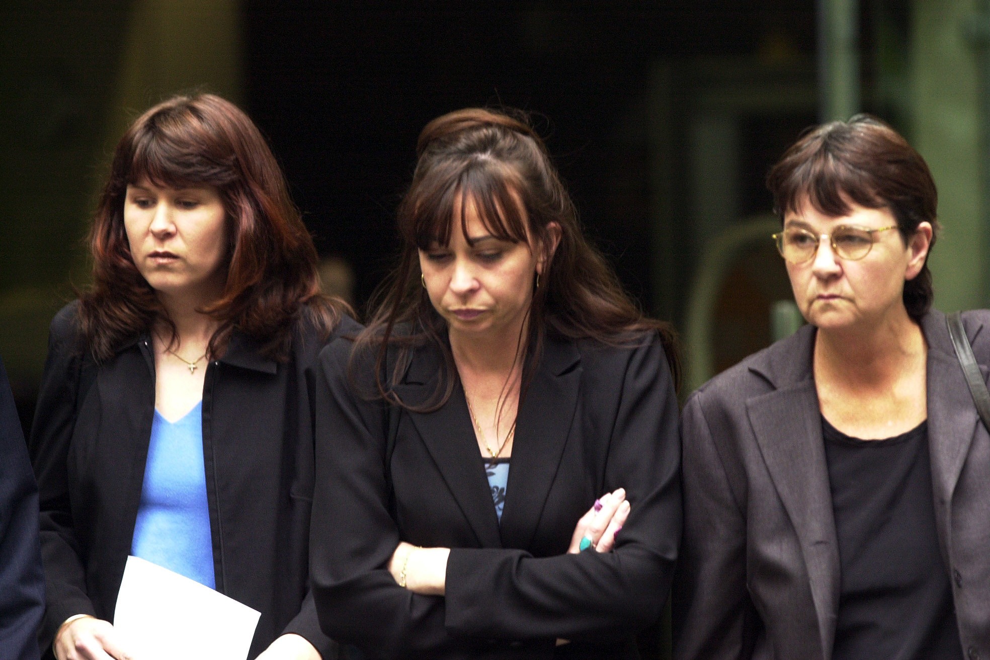 Three women dressed in dark suits walk together with their heads down.