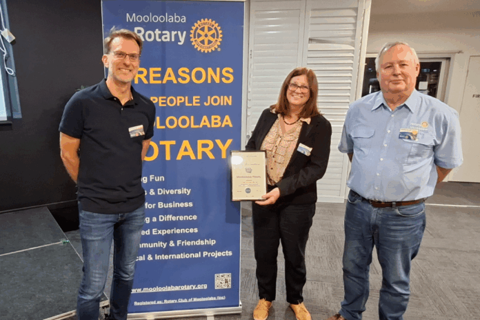 Two men and a woman standing in front of a blue Rotary Club sign. The woman is holding a framed certificate.
