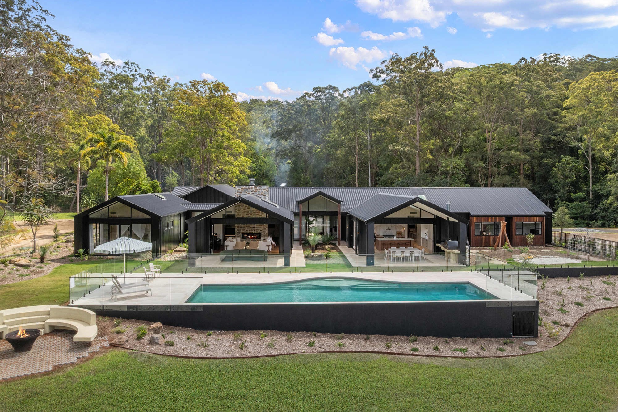 A modern sprawling one-level home with a black roof, overlooking a pool and grassed area, backed by bushland.